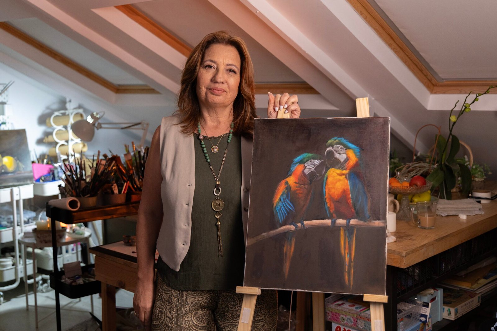 Artist standing next to an easel displaying a painting of two colorful macaws in a studio setting.